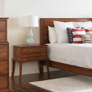 Classic cherry wood bedroom nightstand with two tiers of drawers and gently tapered legs, shown beside a panel bed and styled with a lamp and patriotic pillows for a cozy, timeless look.
