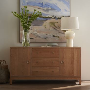 Contemporary solid wood storage consule with brass hardware, topped with a stylish lamp and a vase of fresh greenery, framed by a large abstract artwork