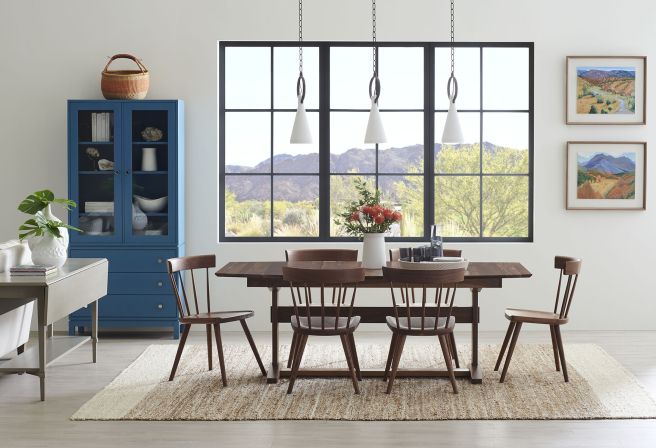 Modern rustic dining space featuring a solid wood trestle table, mid-century spindle chairs, and a bold blue cabinet against a scenic backdrop.