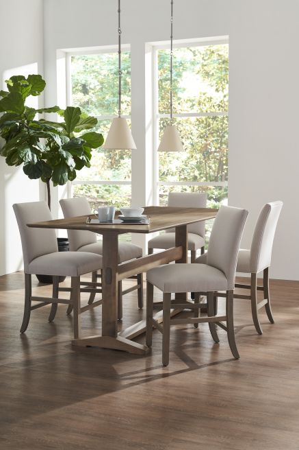 Counter height dining table with a trestle base, surrounded by upholstered chairs in a bright dining area with natural light and modern decor.
