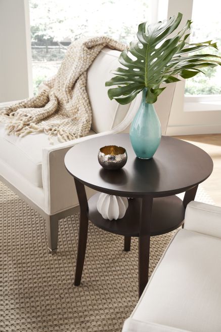 Close-up of a round wooden side table with sleek legs, displaying a teal vase and accent pieces, adjacent to a cushioned armchair