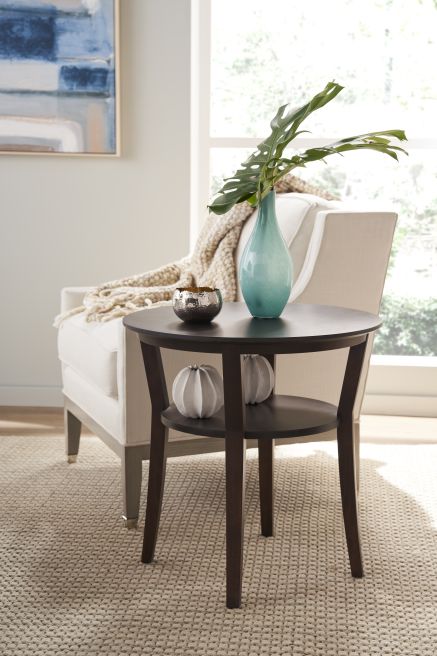 Round wooden side table with a dark finish, supporting a teal vase and decorative items, positioned next to a cream-colored armchair.