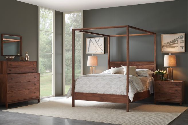 Wood canopy bed with clean lines, paired with a matching nightstand and dresser in a modern bedroom with floor-to-ceiling windows.