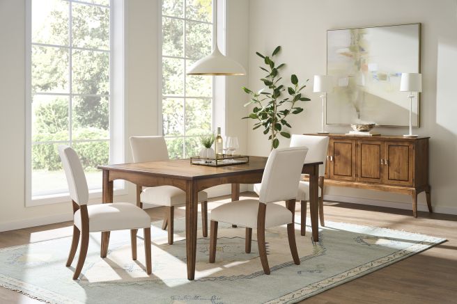 Warm wooden dining table with soft upholstered chairs, set in a bright, airy room with large windows.