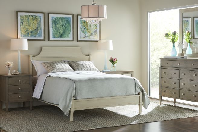 Elegant wooden bed frame with a curved headboard, flanked by matching wood nightstands and a tall dresser, styled with decorative lamps and floral accents.