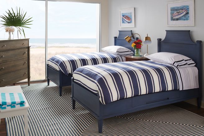 Two navy-blue solid wood twin beds with curved headboards, featuring striped bedding, set in a coastal-themed bedroom with a view of the ocean through a large window.