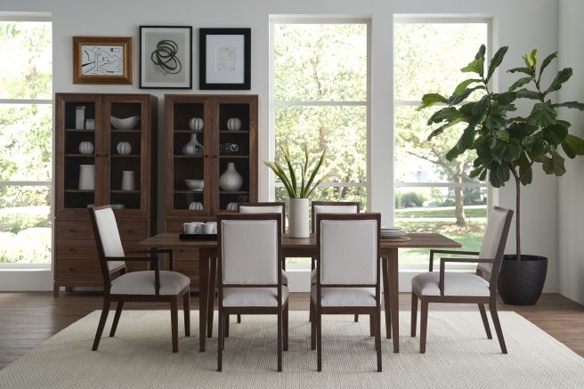 Contemporary dining set featuring a wooden table, white cushioned chairs, two matching walnut display cabinets featuring glass-paneled doors, and large green plant by the window.