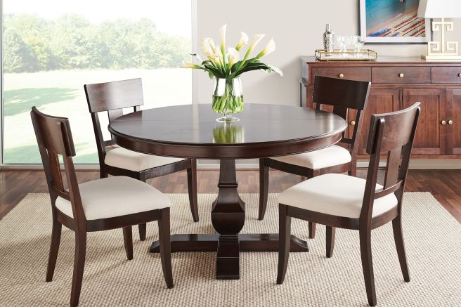 Round pedestal dining table with a dark wood finish, paired with four upholstered chairs, set against a bright window backdrop in a cozy dining room.