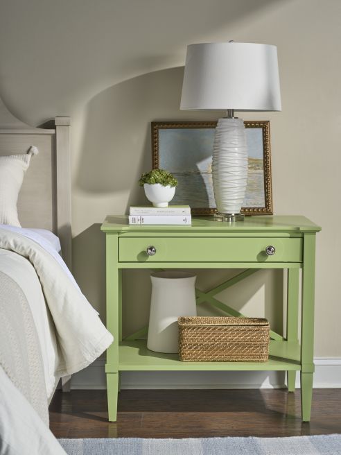 Light green hardwood bedside table with drawer and lower shelf, holding decor pieces next to a neutral upholstered bed.