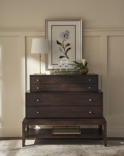 Elegant dark wood chest of drawers with a lower shelf, styled with a table lamp, framed floral artwork, and delicate decor, creating a refined look.