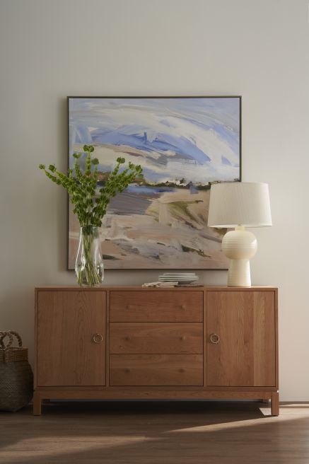 Modern wooden consule with clean lines and brass accents, styled with a table lamp and a tall vase of greenery, set against a large abstract landscape painting.