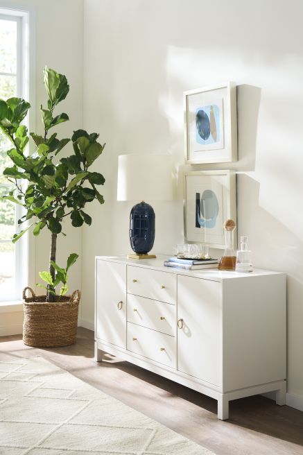 Contemporary wood storage cabinet with brass hardware, complemented by a wicker basket planter and navy blue lamp.