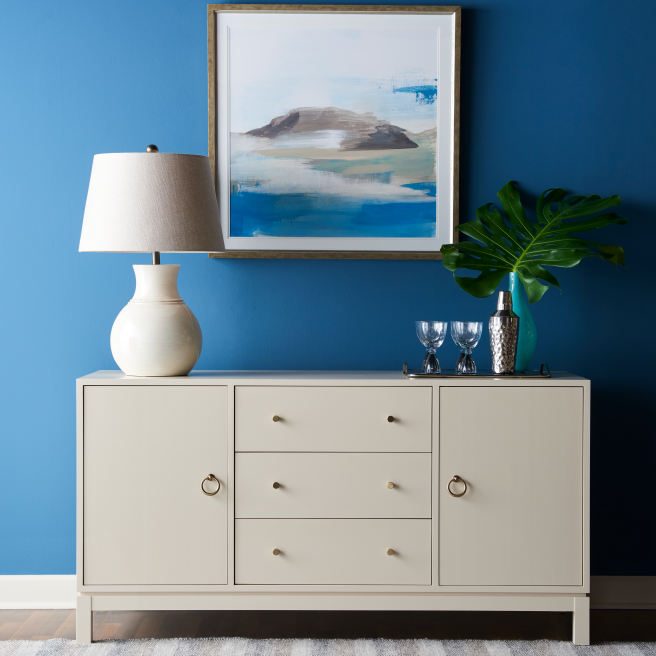 Minimalist consule with clean lines and brass hardware, decorated with a ceramic lamp and a large leaf in a glass vase, framed by an abstract landscape painting on a blue wall.
