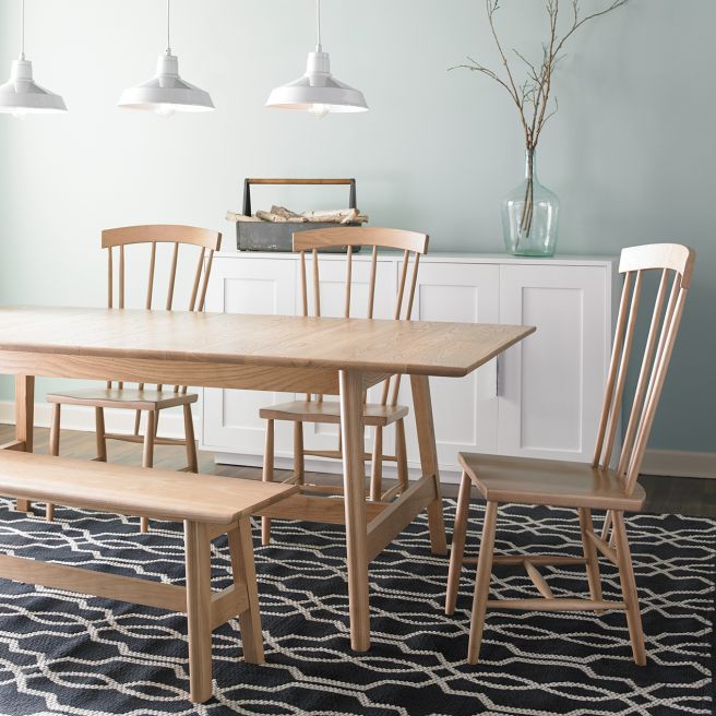 A natural wood dining set featuring a rectangular table, matching bench, and spindle-back chairs in a bright and airy dining room with soft blue walls and modern pendant lighting.
